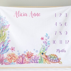 May include: A white baby milestone blanket with a colorful underwater scene. The blanket features a sea turtle, coral, seaweed, and bubbles. The text "Alicia Anne" and "1 2 3 4 5 6 7 8 9 10 11 12 Months" is printed on the blanket.