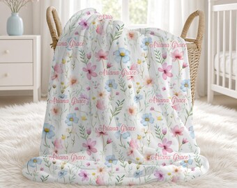 May include: A white blanket featuring a repeating pattern of watercolor-style pink and blue flowers, green leaves, and the name Ariana Grace printed in a pink cursive script throughout the fabric, draped over a woven wicker basket.
