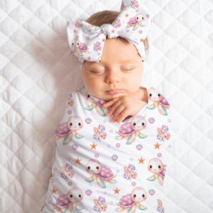 May include: A baby swaddled in a white blanket and wearing a matching headband, both adorned with a repeating pattern of cartoon sea turtles, starfish, and coral in pastel colors. The baby is lying on a white quilted surface.