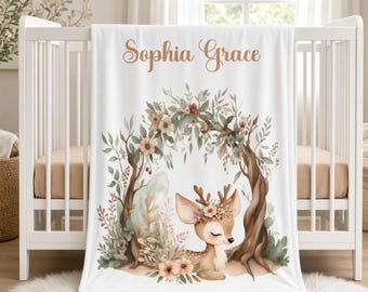 May include: A white baby blanket featuring the name Sophia Grace in brown script text. The design includes a watercolor illustration of a fawn resting under a floral archway with green leaves, brown branches, and soft pink flowers.