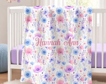 May include: A white baby blanket featuring a watercolor floral pattern in shades of pink, blue, and purple. The name Hannah Ann is printed in a pink cursive script across the center of the blanket, which is draped over a white crib.