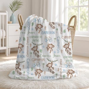 May include: A white fleece blanket featuring a repeating pattern of brown cartoon monkeys, blue balloons, and green palm leaves. The text Cameron and Cameron James is printed in various blue, brown, and green fonts throughout the fabric design.