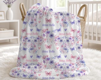May include: A white blanket featuring a repeating pattern of purple and pink watercolor flowers, butterflies, and small hearts. The name Anna Belle is printed in a purple script font throughout the design. The blanket is draped over a wicker basket.