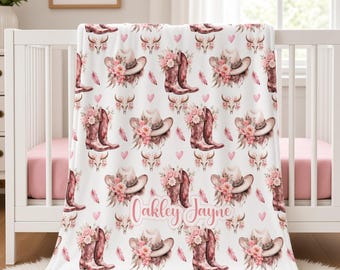 May include: A white fleece blanket featuring a repeating pattern of pink cowboy boots, cowboy hats, floral arrangements, cow skulls, and small hearts. The name Oakley Jayne is printed in a cursive pink script across the centre of the blanket.