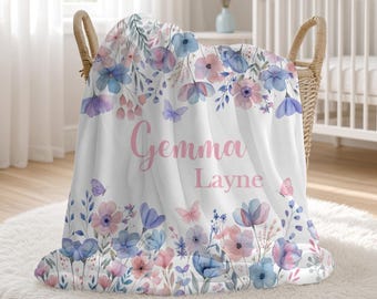 May include: A white blanket featuring a watercolor floral pattern in shades of blue, purple, and pink with small butterflies. The name Gemma Layne is printed in the center in a pink script font. The blanket is draped over a woven basket.