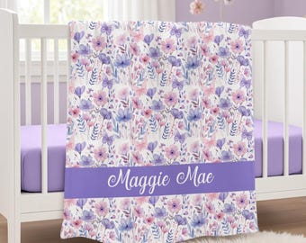 May include: A white baby blanket featuring a repeating pattern of pink and purple flowers, green leaves, and butterflies. A solid purple horizontal band across the lower section displays the name Maggie Mae in white script lettering.
