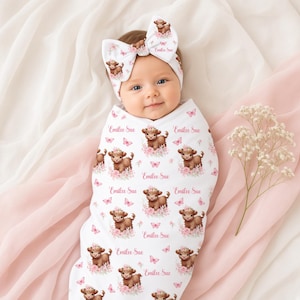May include: A white baby swaddle blanket and matching headband set featuring a repeating pattern of brown highland cows, pink flowers, butterflies, and the name Emilee Sue printed in a cursive pink script across the fabric surface.