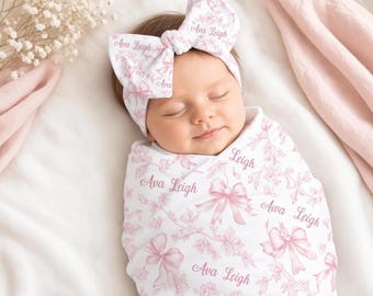 May include: A white swaddle blanket and matching knotted headband featuring a repeating pattern of soft pink bows and floral sprigs, with the name Ava Leigh printed in a cursive script throughout the fabric design.