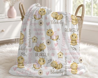 May include: A white fleece blanket featuring a repeating pattern of yellow cartoon bees, pink hearts, small beehives, and the name Blakely Layne printed in various fonts and shades of pink and brown throughout the fabric surface.