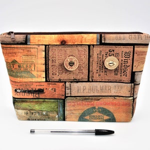 May include: A rectangular cosmetic bag with a vintage wooden crate design. The bag features a zipper closure and is adorned with various text labels and wooden knobs. The bag is made of fabric and has a brown zipper.