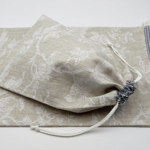 Könnte beinhalten: Eine beigefarbene Leinentasche mit Kordelzug, verziert mit einem weißen Blumenmuster und einem blauen und weißen Gingham-Besatz. Die Tasche ist geschlossen und hat einen weißen Kordelzug.