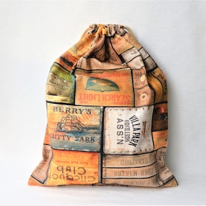 Può includere: Una borsa a cordoncino realizzata in tessuto con un design a patchwork che presenta etichette e illustrazioni in stile vintage. Le etichette includono testo come "Search Light", "Berry's", "Cutty Sark", "Villa Park Orchards Ass'n" e "P. Bulmer Cider Makers".