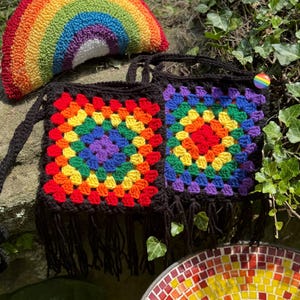May include: A rainbow-themed collection featuring a crochet rainbow pillow, two square crochet bags with fringe, and a mosaic dish. The bags have a black background with colorful squares. A rainbow button is attached to one bag.