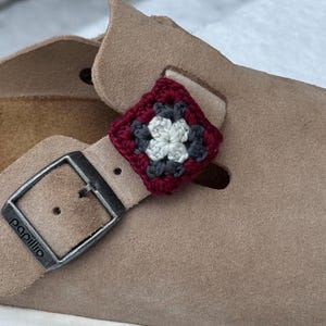 May include: Close-up of a tan suede sandal with a silver buckle and a crocheted square charm. The charm features a floral design in red, gray, and white. The sandal is on a white sole.