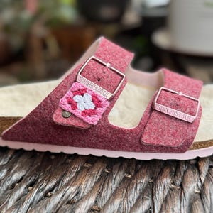 May include: Close-up of a pair of Birkenstock sandals in a deep rose color. The sandals feature a crochet embellishment with pink and white floral details. The buckles are silver, and the footbed is a light tan color.