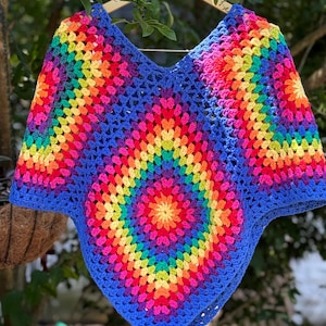 May include: A crochet poncho featuring a vibrant granny square pattern with concentric rings of rainbow colors including red, orange, yellow, green, blue, and purple, all framed by a solid blue yarn border with a V-neckline and pointed hem.