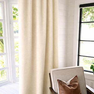 May include: Cream-colored linen curtains hang from a black rod in front of a window. A light-colored armchair with a brown leather pillow sits nearby. The room has white walls and a window seat.