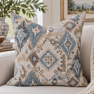 May include: A square throw pillow featuring a woven fabric with a multi-colored geometric pattern in shades of light blue, dark blue, beige, and brown. The design consists of repeating diamond and angular shapes across the entire surface.