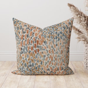 May include: A square cushion featuring a repeating pattern of abstract shapes in orange, blue, and brown on a cream background. The cushion is placed on a wooden floor.