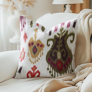 May include: A white throw pillow with a colorful ikat pattern in shades of red, green, yellow, and purple. The pillow is sitting on a beige couch.