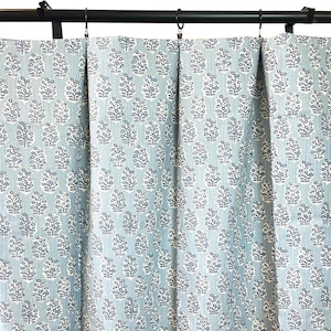 May include: Light blue and white floral patterned curtains hanging from a black rod with rings. The curtains have a repeating pattern of stylized floral motifs in white against a light blue background. The fabric is gathered in soft folds.