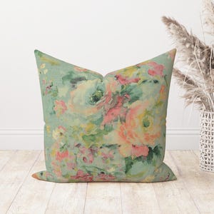 Op de afbeelding: Een decoratief kussen met een bloemenpatroon in tinten roze, geel en groen op een lichtblauwe achtergrond.