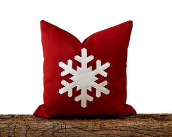May include: A red square pillow with a large white snowflake design. The pillow is resting on a rustic wooden surface. The snowflake is centered on the pillow.