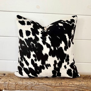 May include: A square decorative pillow with a cowhide pattern. The pillow features a black and white spotted design, resembling a cow's hide. The pillow is placed on a rustic wooden bench, creating a farmhouse aesthetic.