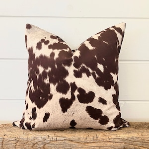 May include: A square decorative pillow with a cowhide pattern. The pillow features a brown and white spotted design, resembling a cow's hide. The pillow is placed on a wooden surface, against a white background.