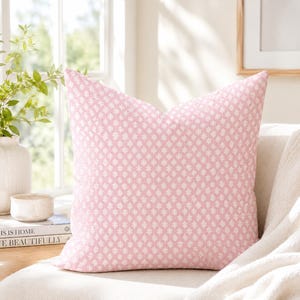 May include: A square throw pillow featuring a soft pink fabric base with a repeating white geometric floral pattern. The pillow has a clean, structured shape with crisp edges and a consistent, evenly distributed print across the entire front surface.