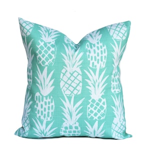 May include: Square decorative pillow with a teal background and a white pineapple pattern. The pillow has a soft, textured appearance and is designed for home decor. The pineapple design is repeated across the entire surface of the pillow.