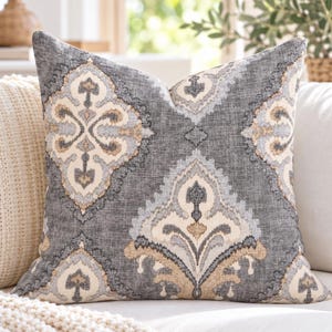 May include: A square throw pillow featuring a dark gray fabric base with a repeating damask pattern in shades of cream, light blue, and tan. The textile has a visible woven texture, and the design consists of symmetrical, ornate shapes.