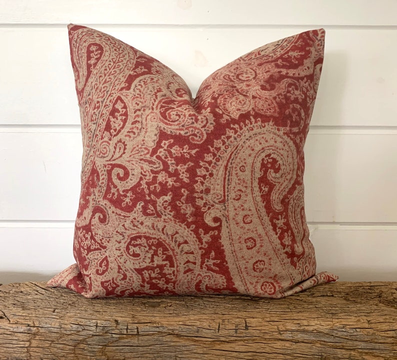 May include: Red and cream paisley patterned throw pillow. The pillow is made of a soft, textured fabric.