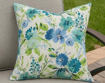 May include: Square throw pillow with a floral pattern in shades of blue and green on a cream background. The pillow is placed on a gray outdoor chair, suggesting a garden or patio setting. The design features watercolor-style flowers and foliage.