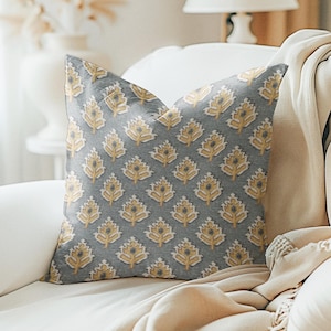 May include: A grey throw pillow with a yellow and white floral pattern. The pillow is sitting on a white sofa with a beige throw blanket.