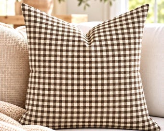May include: A square throw pillow featuring a classic brown and white gingham check pattern. The fabric displays a consistent grid of small, alternating dark brown and off-white squares across the entire surface of the cushion cover.