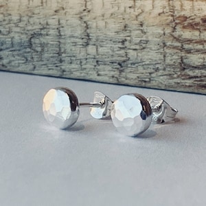 Puede incluir: Un par de pendientes de botón de plata martillados. Los pendientes son redondos y tienen una superficie texturizada.