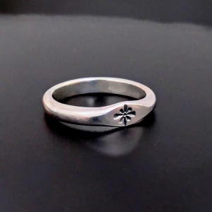 May include: A silver ring with a floral design. The ring is made of a shiny metal and features a small, detailed flower emblem on the front. The band is smooth and rounded, creating a simple yet elegant look. The ring is shown against a dark background.