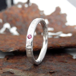 May include: A silver ring with a textured finish, featuring a small, round, pink gemstone. The ring is displayed against a rusty brown backdrop, accentuating the contrast between the silver and the warm hues.