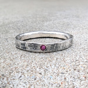 May include: A silver band ring featuring a textured, grid-like pattern across its surface. The ring is accented with a single, small, round, vibrant pink gemstone set flush into the centre of the band, creating a distinct contrast against the metal.