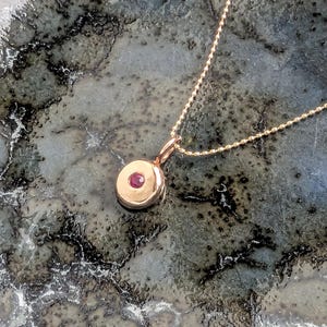 May include: A rose gold necklace featuring a delicate ball chain and a small, circular pendant. The pendant has a smooth, polished finish and is set with a single, round, vibrant pink gemstone at its centre, creating a refined and simple look.