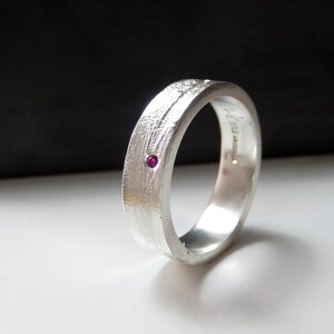 May include: A silver band with a textured finish and a small ruby gemstone set into the surface.