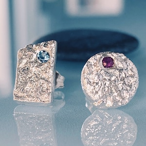 May include: A pair of silver earrings with textured surfaces. One earring features a blue gemstone, while the other has a pink gemstone.
