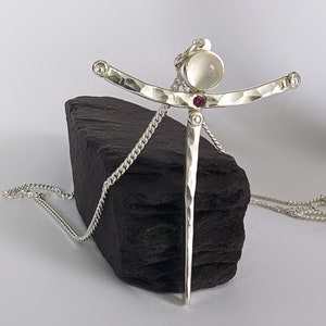 May include: A silver necklace with a pendant featuring a hammered silver design with a white moonstone and two small pink gemstones. The necklace is on a black surface.