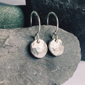 Può includere: Un paio di orecchini pendenti in argento con ciondoli rotondi martellati. Gli orecchini sono appesi a una roccia grigia.