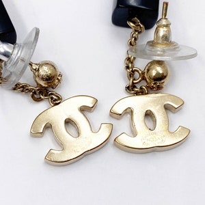 May include: A pair of gold-tone Chanel earrings with a black and gold chain and a CC logo charm. The earrings are made of metal and have a post back.