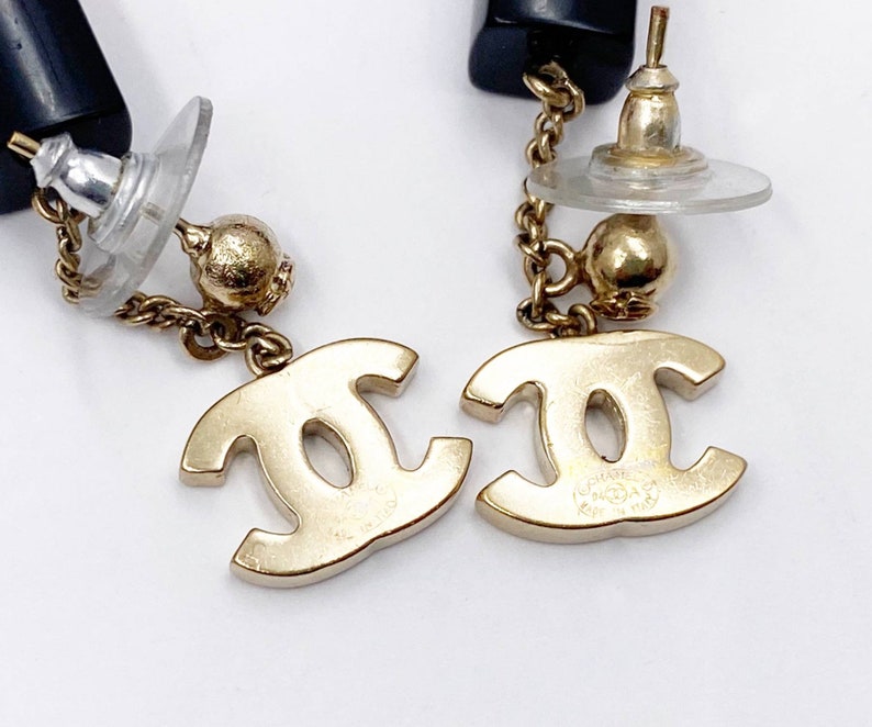May include: A pair of gold-tone Chanel earrings with a black and gold chain and a CC logo charm. The earrings are made of metal and have a post back.