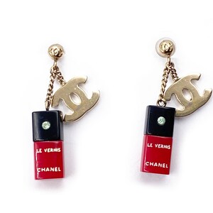 May include: A pair of gold tone earrings with a black and red nail polish bottle charm and a double 'C' logo charm. The charms hang from a gold chain.