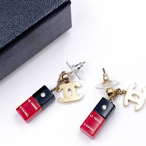 May include: A pair of gold tone Chanel earrings with red and black enamel charms that say "Chanel" and "Le Vernis".