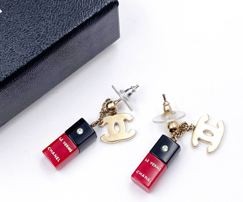 May include: A pair of gold tone Chanel earrings with red and black enamel charms that say "Chanel" and "Le Vernis".